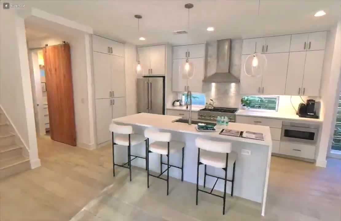 A still from a Gaussian splat walkthrough of a real kitchen, rendered in a browser tab. Light wood floor, white island with three bar stools, pendant lights, and a SuperSplat watermark in the top-left corner showing this is a live 3D scene, not a photo