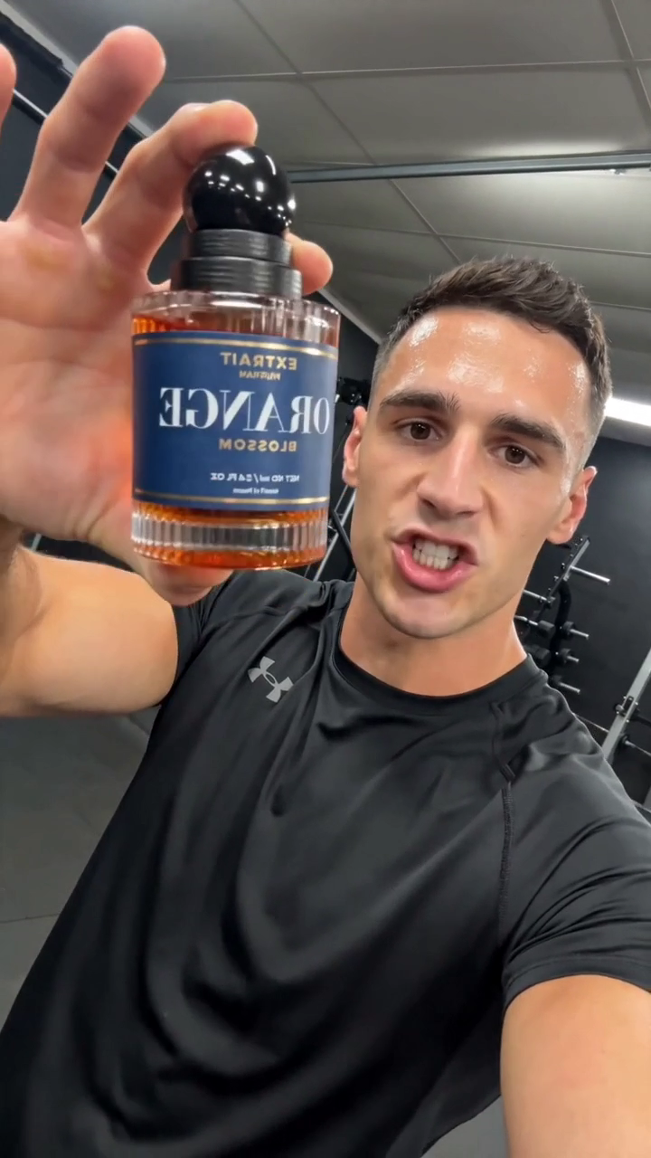 A synthetic 27-year-old man in a black athletic tee in a home gym, holding a cologne bottle up to a selfie-style phone camera